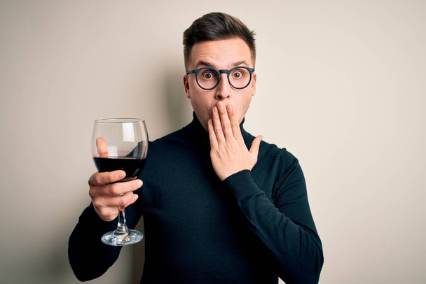 Giovane uomo caucasico bello bere un bicchiere alcolico di vino rosso su fondo isolato coprire la bocca con la mano scioccata dalla vergogna per errore, espressione di paura, paura in silenzio, concetto segreto
 - Foto, immagini