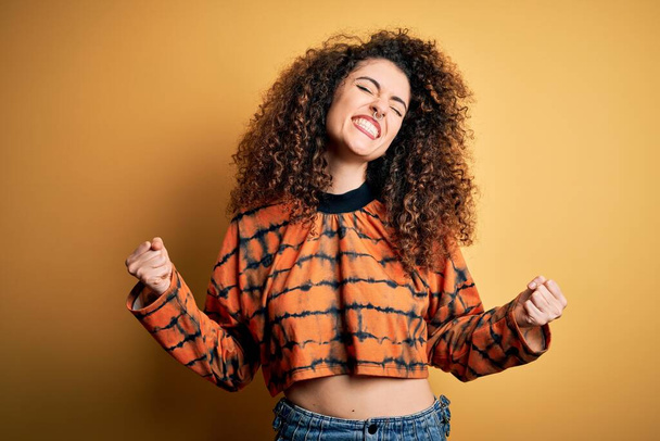 Young beautiful brunette woman with curly hair and piercing wearing casual t-shirt very happy and excited doing winner gesture with arms raised, smiling and screaming for success. Celebration concept. - Photo, image