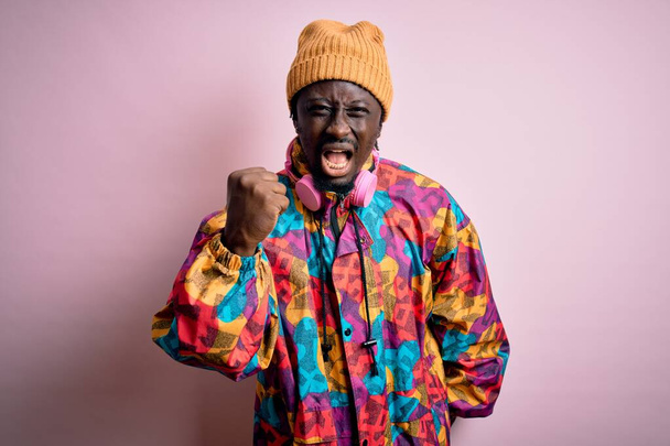 Young handsome african american man wearing colorful coat and cap over pink background angry and mad raising fist frustrated and furious while shouting with anger. Rage and aggressive concept. - Photo, Image