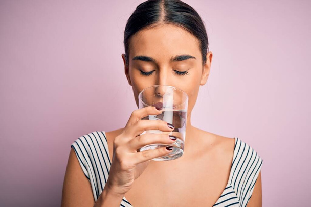 Молода красива брюнетка п'є склянку здорової води для відпочинку, стоячи над ізольованим рожевим тлом
 - Фото, зображення