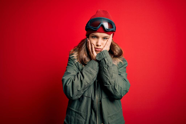 Νεαρό ξανθό κορίτσι που φοράει γυαλιά σκι και χειμερινό παλτό για τον καιρό του σκι πάνω από το κόκκινο φόντο Κουρασμένα χέρια που καλύπτουν το πρόσωπο, την κατάθλιψη και τη θλίψη, αναστατωμένος και ερεθισμένος για το πρόβλημα - Φωτογραφία, εικόνα