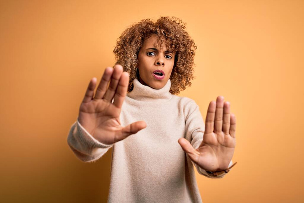 Giovane bella donna afro-americana indossa maglione collo alto su sfondo giallo facendo gesto stop con le mani palme, espressione arrabbiata e frustrazione
 - Foto, immagini