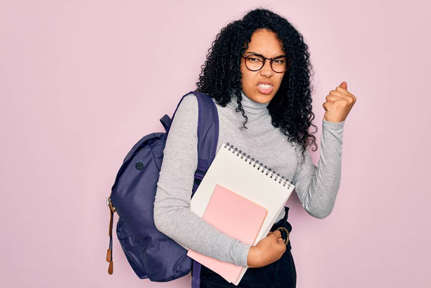 Young african american curly student woman wearing backpack and glasses holding book annoyed and frustrated shouting with anger, crazy and yelling with raised hand, anger concept - Foto, Imagem