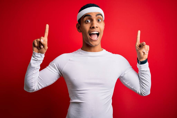 Νεαρός όμορφος Αφροαμερικάνος αθλητής που φοράει αθλητικά ρούχα πάνω από κόκκινο φόντο χαμογελώντας έκπληκτος και δείχνοντας με τα δάχτυλα και σήκωσε τα χέρια. - Φωτογραφία, εικόνα