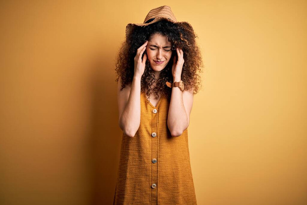 Beautiful brunette woman on vacation with curly hair and piercing wearing hat and dress suffering from headache desperate and stressed because pain and migraine. Hands on head. - Фото, изображение