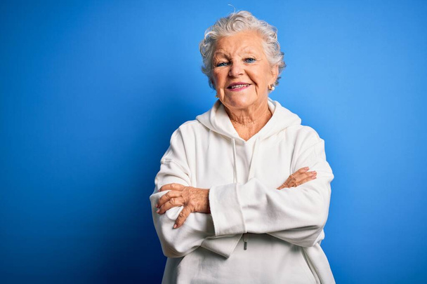 Senior beautiful sporty woman wearing white sweatshirt over isolated blue background happy face smiling with crossed arms looking at the camera. Positive person. - Photo, Image