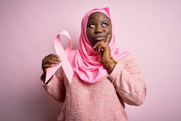 Jonge Afro-Amerikaanse plus size vrouw draagt moslim hijab met roze kankerlint serieus gezicht denken over vraag, zeer verward idee - Foto, afbeelding
