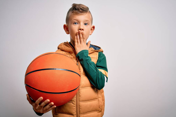 Νεαρό μικρό καυκάσιο αθλητικό παιδί που παίζει μπάσκετ κρατώντας πορτοκαλί μπάλα πάνω από απομονωμένο φόντο καλύπτουν το στόμα με το χέρι συγκλονισμένος από ντροπή για λάθος, έκφραση του φόβου, φοβάται στη σιωπή, μυστική έννοια - Φωτογραφία, εικόνα