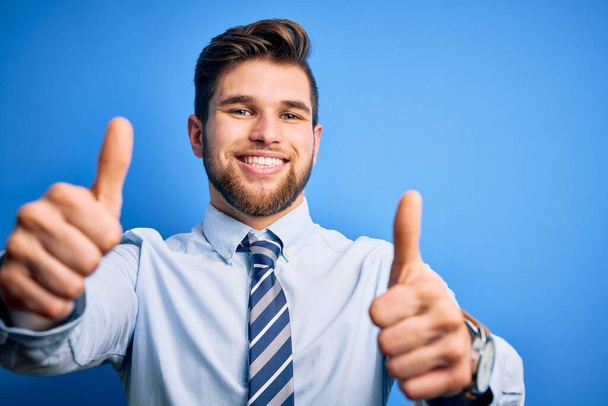 ひげと青い目を持つ若いブロンドのビジネスマンはエレガントなシャツを着て、手で肯定的なジェスチャーを行うことを承認立ってネクタイ、親指を笑顔と成功のために幸せ。勝者のジェスチャー. - 写真・画像