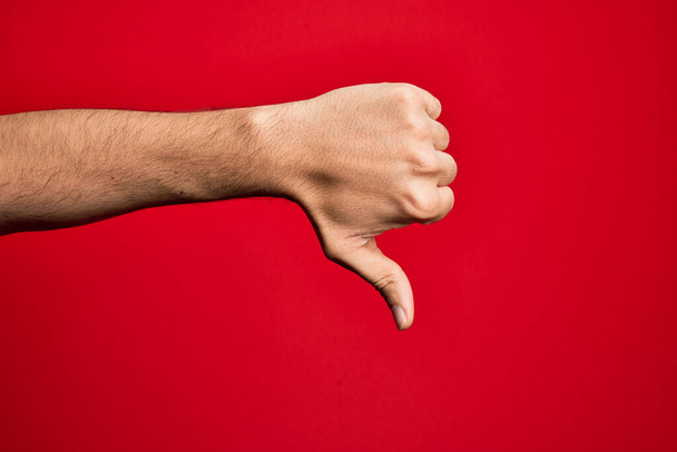 Рука кавказского молодого человека показывает пальцы на изолированном красном фоне, делая большие пальцы вниз жест отказа, неодобрение неприязни и негативный знак - Фото, изображение