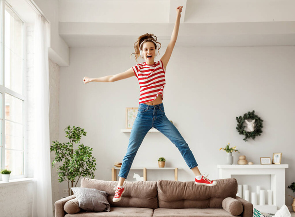 Excited young female in wireless headphones and casual clothes enjoying life while listening to music and jumping and dancing alone of sofa in cozy light apartmen - Foto, Imagem