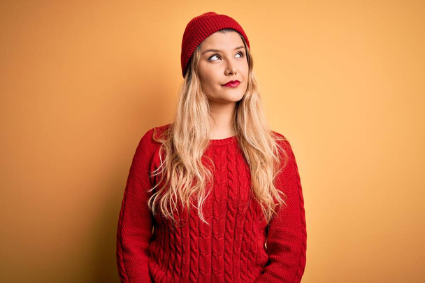 Young beautiful blonde woman wearing casual sweater and wool cap over white background smiling looking to the side and staring away thinking. - Photo, Image