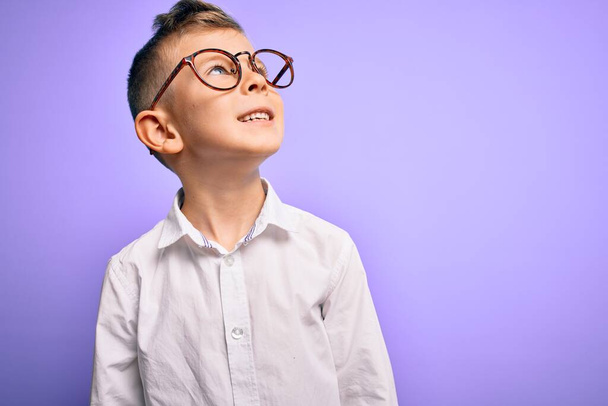 Young little caucasian kid with blue eyes wearing glasses and white shirt over purple background looking away to side with smile on face, natural expression. Laughing confident. - Foto, immagini