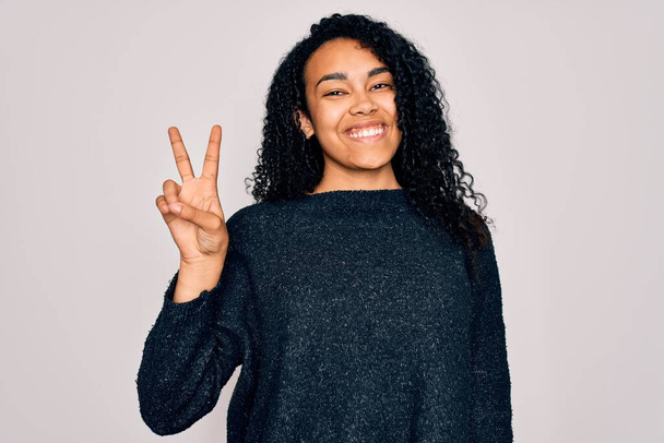 Young african american curly woman wearing casual sweater standing over white background smiling with happy face winking at the camera doing victory sign. Number two. - Photo, Image