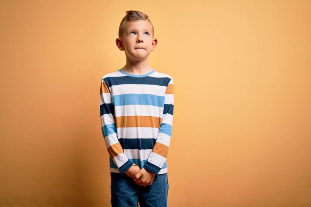 Маленький белый мальчик с голубыми глазами в разноцветной полосатой рубашке на жёлтом фоне улыбается и смотрит в сторону, думая:
. - Фото, изображение