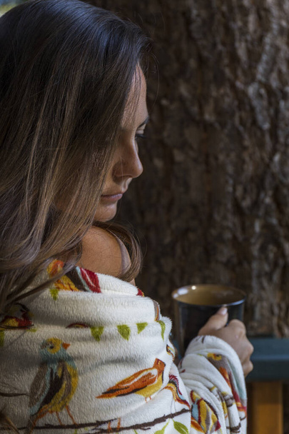 tummahiuksinen nainen, jolla on löysät hiukset, käärittynä huopaan juo kahvia aamulla
 - Valokuva, kuva