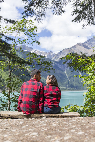 couple dressed alike, sitting on a wooden bench looking at each other, in the background you can see the turquoise lake and the mountains - Photo, Image