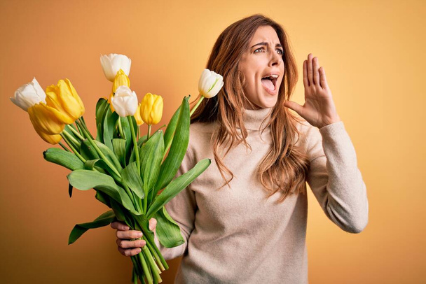 Giovane bella donna bruna che tiene bouquet di tulipani gialli su sfondo isolato urlando e urlando forte a lato con mano sulla bocca. Concetto di comunicazione
. - Foto, immagini