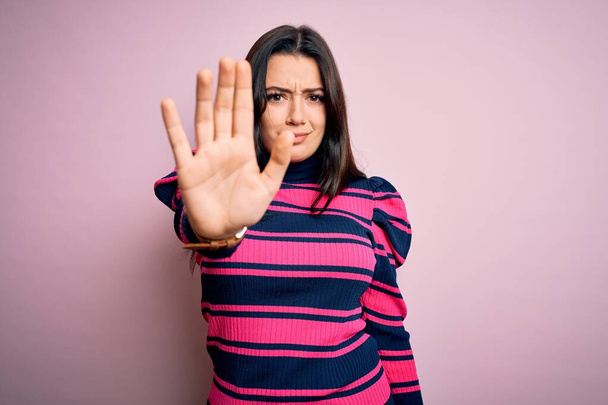 Jeune femme brune élégante portant une chemise rayée sur fond rose isolé faisant arrêter de chanter avec la paume de la main. Expression d'avertissement avec geste négatif et sérieux sur le visage
. - Photo, image
