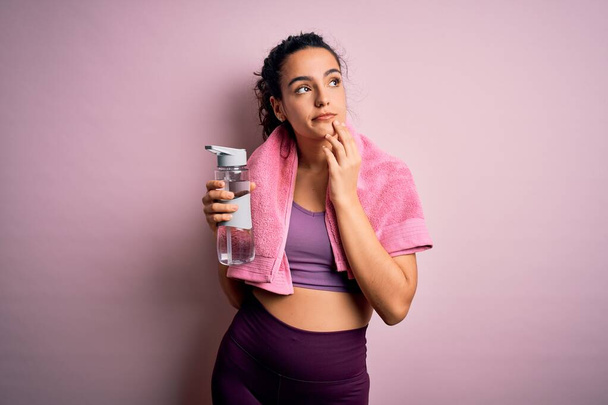 Young beautiful sportswoman with curly hair doing sport drinking bottle of water using towel serious face thinking about question, very confused idea - Photo, Image