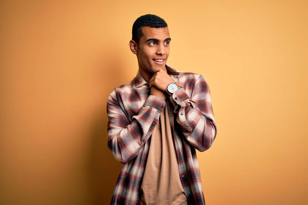 Young handsome african american man wearing casual shirt standing over yellow background laughing nervous and excited with hands on chin looking to the side - Photo, Image