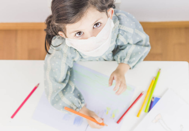 Маленька дівчинка вчиться і малює вдома або в школі з маскою. Краєвид. Коронавірус. Коїд 19 - Фото, зображення