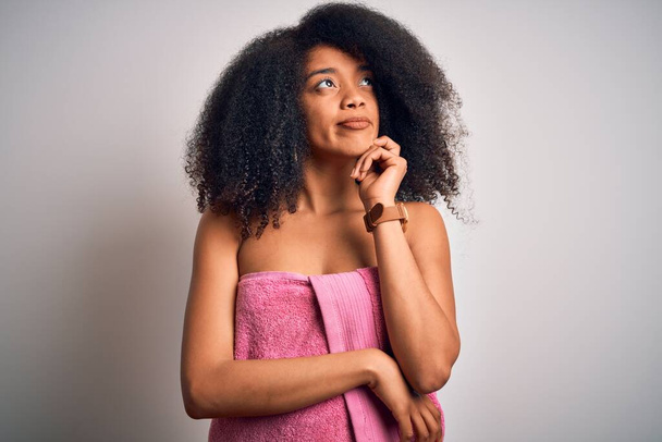 Fiatal afro-amerikai nő afro hajjal, törülközőt visel a szépségápolási zuhany után, kézzel az állán, gondolkodva a kérdésen, elmélkedő arckifejezéssel. Gondos arccal mosolygott. Kétséges fogalom. - Fotó, kép