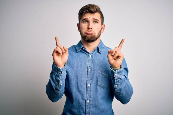 Young handsome blond man with beard and blue eyes wearing casual denim shirt Pointing up looking sad and upset, indicating direction with fingers, unhappy and depressed. - Photo, Image
