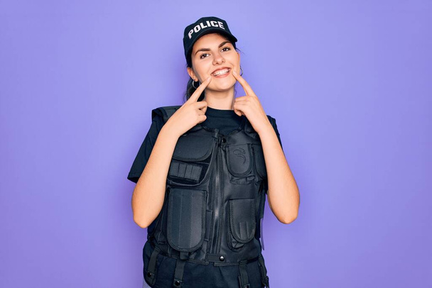 Молодая полицейская женщина в пуленепробиваемой форме на фиолетовом фоне улыбается с открытым ртом, указывает пальцами и заставляет веселую улыбку
 - Фото, изображение