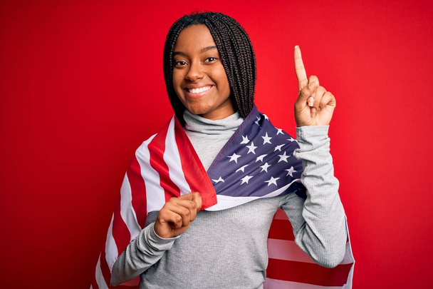 Junge afrikanisch-amerikanische Patriotin, die am Unabhängigkeitstag die Flagge der Vereinigten Staaten von Amerika trägt, überrascht mit einer Idee oder Frage, die mit glücklichem Gesicht mit dem Finger zeigt, Nummer eins - Foto, Bild