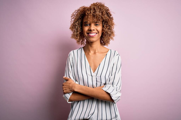Beautiful african american woman with curly hair wearing striped t-shirt over pink background happy face smiling with crossed arms looking at the camera. Positive person. - Photo, Image
