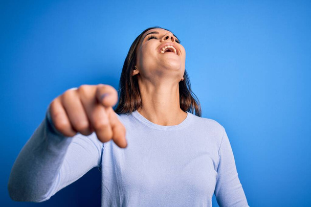 Young beautiful brunette woman wearing casual sweater standing over blue background laughing at you, pointing finger to the camera with hand over body, shame expression - Photo, Image