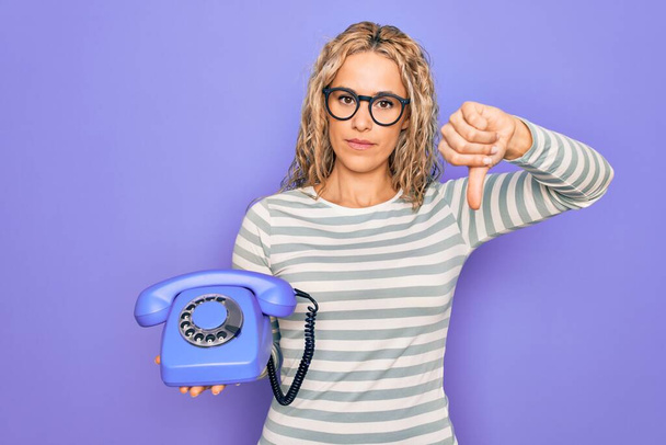 Молодая красивая блондинка держит винтажный телефон на изолированном фиолетовом фоне с сердитым лицом, негативный знак показывает неприязнь с опущенными пальцами, отвержение концепции
 - Фото, изображение