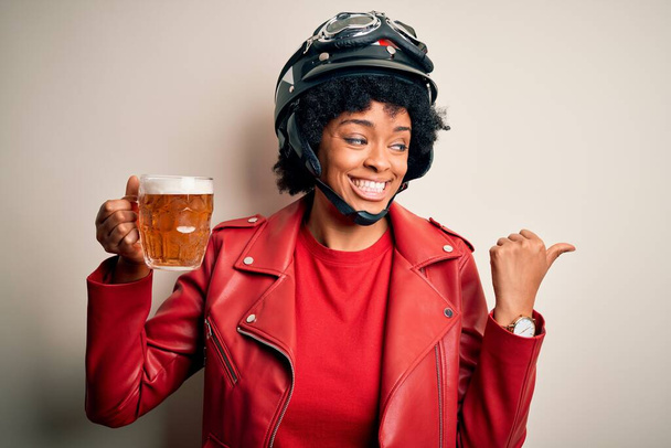 Jovem afro-americana afro motociclista com cabelo encaracolado bebendo jarra de cerveja apontando e mostrando com o polegar até o lado com o rosto feliz sorrindo
 - Foto, Imagem