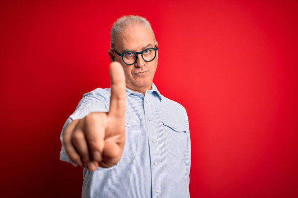Ένας μεσήλικας όμορφος άντρας, με ριγέ πουκάμισο και γυαλιά πάνω από κόκκινο φόντο, δείχνει με το δάχτυλο ψηλά και θυμωμένη έκφραση, χωρίς να δείχνει καμία χειρονομία. - Φωτογραφία, εικόνα