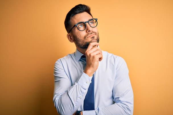 Young handsome businessman wearing tie and glasses standing over yellow background with hand on chin thinking about question, pensive expression. Smiling with thoughtful face. Doubt concept. - Photo, Image