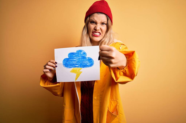 Young beautiful blonde woman wearing raincoat holding banner with cloud thunder image annoyed and frustrated shouting with anger, crazy and yelling with raised hand, anger concept - Photo, Image