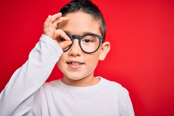 Młody, mądry chłopiec nosi okulary kujonów na czerwonym, odizolowanym tle z uśmiechniętą twarzą robi znak ok z ręką na oku patrząc przez palce - Zdjęcie, obraz