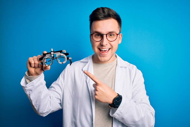 Joven hombre óptico profesional guapo sosteniendo gafas optométricas sobre fondo azul muy feliz señalando con la mano y el dedo
 - Foto, Imagen