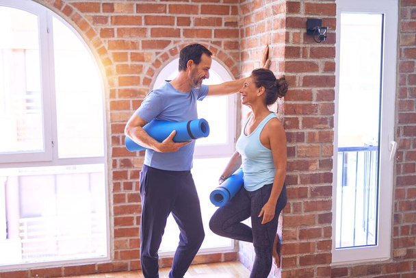 Middle age beautiful sporty couple smiling happy. Standing with smile on face holding mat and speaking at gym - Fotoğraf, Görsel