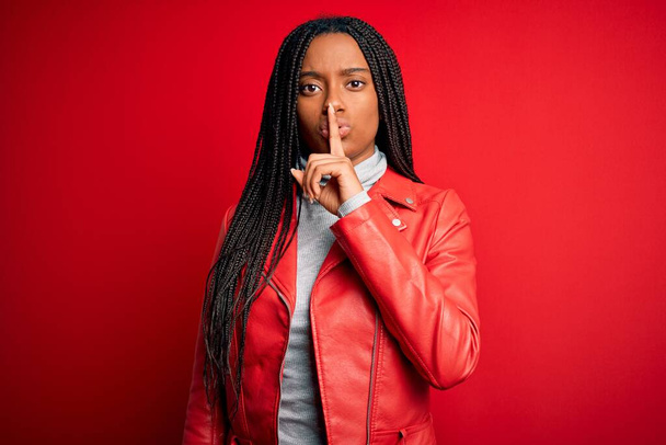 Jonge Afro-Amerikaanse vrouw draagt coole mode leren jas over rood geïsoleerde achtergrond vragen om stil te zijn met de vinger op de lippen. Stilte en geheim concept. - Foto, afbeelding