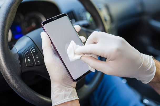 Close up of driver hands in surgical gloves wiping mobile phone with antibacterial wet wipe inside of car. - Fotó, kép