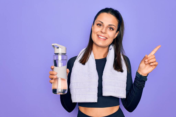 Молодая красивая брюнетка спортивная женщина с помощью спортивного полотенца пить бутылку воды улыбаясь счастливый указывая рукой и пальцем в сторону
 - Фото, изображение