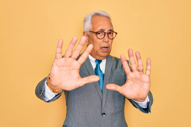 Hombre de negocios guapo de pelo gris de mediana edad con gafas sobre fondo amarillo haciendo gesto de stop con palmas de las manos, expresión de enojo y frustración
 - Foto, imagen