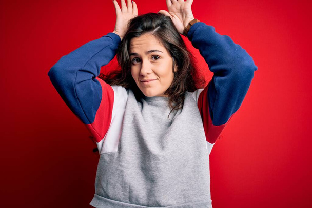 Young beautiful woman with curly hair wearing casual sweatshirt over isolated red background Doing bunny ears gesture with hands palms looking cynical and skeptical. Easter rabbit concept. - Photo, Image