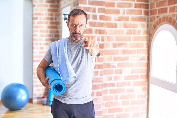 Middle age handsome sportman holding mat and towel standing before do exercise at gym pointing with finger to the camera and to you, hand sign, positive and confident gesture from the front - Zdjęcie, obraz