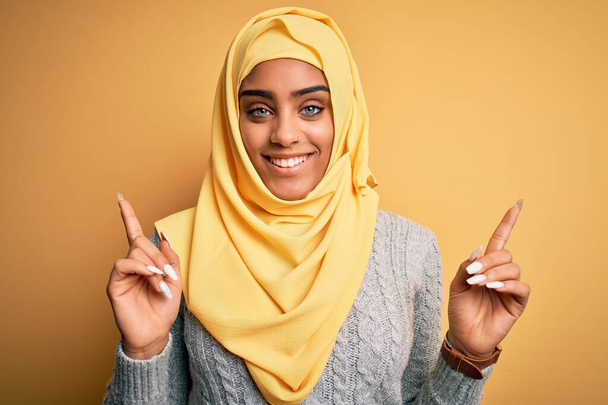 Giovane bella ragazza afro-americana che indossa hijab musulmano su sfondo giallo isolato sorridente fiducioso punta con le dita in direzioni diverse. Copia spazio per pubblicità
 - Foto, immagini