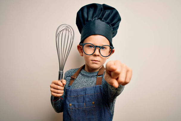 Junge kleine kaukasische Koch-Kind trägt Kochuniform und Hut mit manuellem Schneebesen zeigt mit dem Finger auf die Kamera und auf Sie, Handzeichen, positive und selbstbewusste Geste von vorne - Foto, Bild