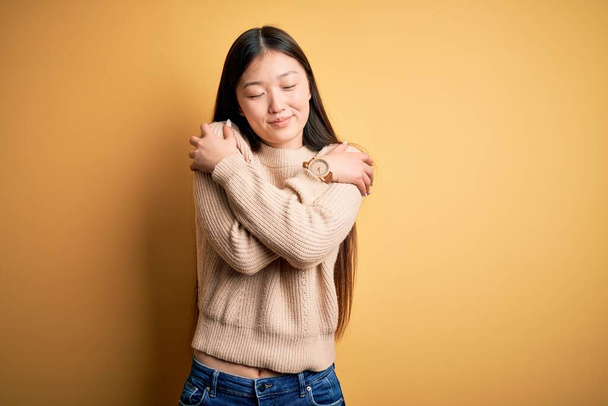 Mladá krásná asijská žena na sobě ležérní svetr přes žluté izolované pozadí objímající se šťastný a pozitivní, usmívající se sebevědomě. Sebeláska a sebepéče - Fotografie, Obrázek