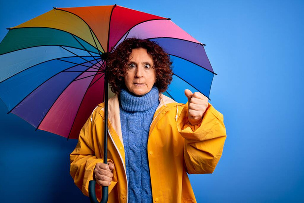 Kobieta w średnim wieku kręcone włosy w płaszczu przeciwdeszczowym trzymając kolorowy parasol na niebieskim tle irytowany i sfrustrowany krzycząc z gniewu, szalony i krzycząc z podniesionymi rękami, koncepcja złości - Zdjęcie, obraz